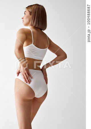 One slender young woman with sportive body shape in cotton inner wear posing over white background. Back view One slender young woman with sportive body shape in cotton inner wear posing over white background. Back view 100488687