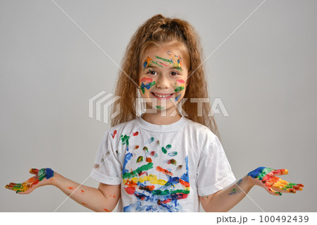 Little girl in white t-shirt is posing standing isolated on white and gesticulating with her painted in different colors palms. Art studio. Close-up. Little girl in white t-shirt is posing standing isolated on white and gesticulating with her painted in different colors palms. Art studio. Close-up. 100492439