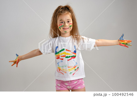 Little girl in white painted t-shirt is posing standing isolated on white and gesticulating with her colored in different paints palms and face. Art studio. Close-up. 100492546