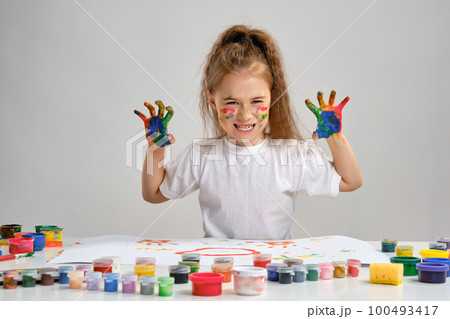 Little girl in white t-shirt sitting at table with whatman and paints on it, posing with painted face and hands. Isolated on white. Medium close-up. 100493417
