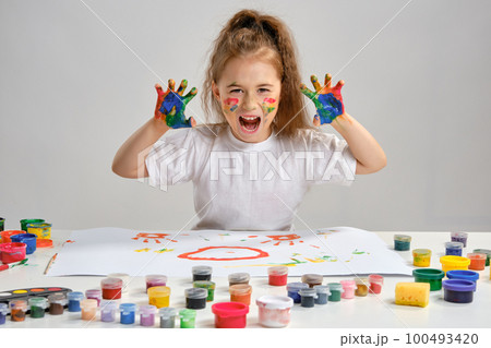 Little girl in white t-shirt sitting at table with whatman and paints on it, posing with painted face and hands. Isolated on white. Medium close-up. Little girl in white t-shirt sitting at table with whatman and paints on it, posing with painted face and hands. Isolated on white. Medium close-up. 100493420