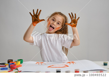 Little girl in white t-shirt sitting at table with whatman and colorful paints, showing her painted hands. Isolated on white. Medium close-up. 100493522