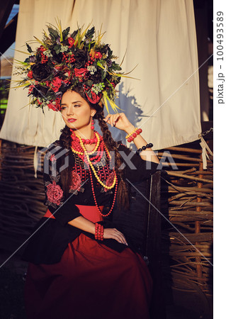 Brunette girl in a black and red embroidered ukrainian authentic national costume and a wreath of flowers is posing against a terrace. Close-up. 100493589