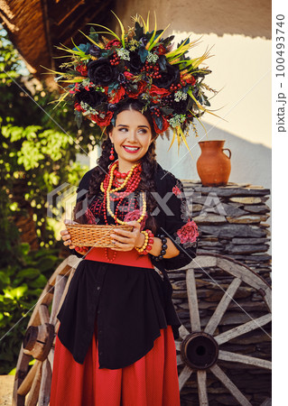 Brunette girl in black and red embroidered ukrainian authentic national costume and a wreath of flowers is posing standing against a white hut. Brunette girl in black and red embroidered ukrainian authentic national costume and a wreath of flowers is posing standing against a white hut. 100493740