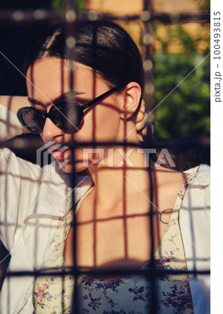 Portrait of girl in sunglasses posing in city behind a trellised fence. Dressed in top with floral print, white shirt, black trousers, waist bag. 100493815