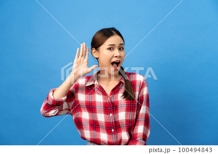 Young asian woman wearing gingham red shirt with braid hairstyle and excited face while use hand to listening Young asian woman wearing gingham red shirt with braid hairstyle and excited face while use hand to listening 100494843