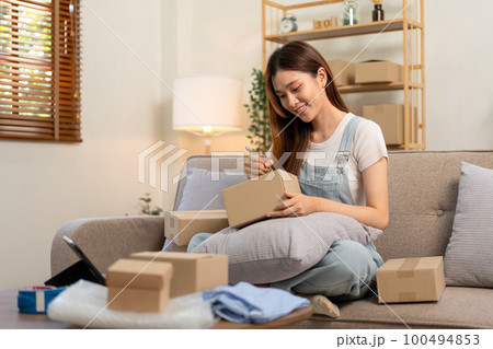 Young entrepreneur is using tablet to checking online orders and writing customer address on cardboard boxes 100494853