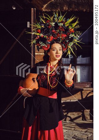 Brunette girl in a black and red ukrainian embroidered authentic national costume and a wreath of flowers is posing standing at the gate. 100495172