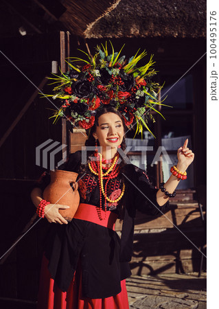 Brunette girl in a black and red ukrainian embroidered authentic national costume and a wreath of flowers is posing standing at the gate. Brunette girl in a black and red ukrainian embroidered authentic national costume and a wreath of flowers is posing standing at the gate. 100495176
