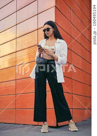 Girl in sunglasses posing in city against an orange building. Dressed in top with floral print, white shirt, black trousers, waist bag and sneakers. 100495194
