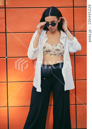 Portrait of girl in dark sunglasses posing in city against orange building. Dressed in top with floral print, white shirt, black trousers, waist bag. 100495226