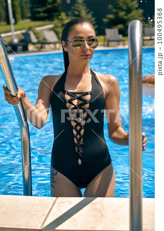 Portrait of a female having rest and posing in a swimming pool. Dressed in a black swimsuit and sunglasses. 100495368