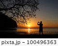 Man taking picture of sunset, beautiful red sky with the shadow of a big tree on the beach 100496363