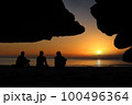 A group of Asian men sitting and looking at the sunset, beautiful red sky. In front of a cave on the beach 100496364