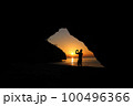 Man taking picture of sunset, beautiful red sky In front of a cave on the beach 100496366
