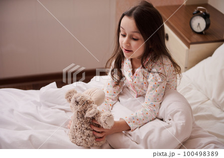 Adorable Caucasian little child girl with beautiful long hair, dressed in stylish pajamas, sitting on the bed in her bedroom and playing with her plush toy sheep, before going to bed. Kids. Childhood 100498389