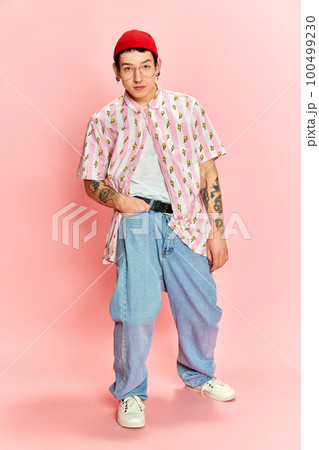Portrait of stylish guy with tattoo keeping hand in pocket and looking at camera over pink background. 100499230