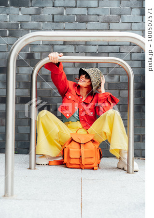 Hipster fashion young woman in bright clothes, sun glasses and bucket hat sitting near gray brick wall background with bike stands frame. Urban city street fashion. Vertical card. Selective focus. Hipster fashion young woman in bright clothes, sun glasses and bucket hat sitting near gray brick wall background with bike stands frame. Urban city street fashion. Vertical card. Selective focus. 100502571