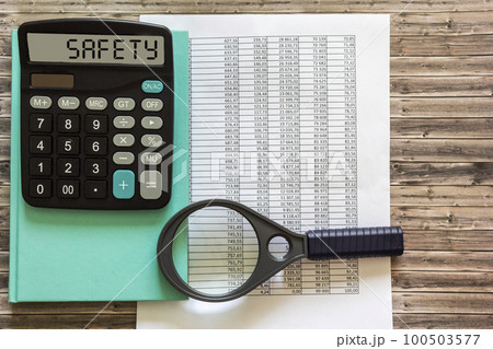 Safety text on calculator display, green notepad, magnifier and financial reports on a wooden table. 100503577