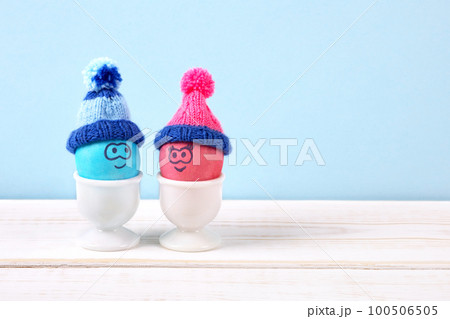 Two Easter eggs with funny faces and crocheted hats in white egg stands on a wooden table. Happy Easter concept. Greeting card. Two Easter eggs with funny faces and crocheted hats in white egg stands on a wooden table. Happy Easter concept. Greeting card. 100506505