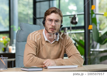 Portrait of a young businessman man in a headset working concentratedly from a home office. Speaks expressively and seriously to the camera, communicates via video call. Portrait of a young businessman man in a headset working concentratedly from a home office. Speaks expressively and seriously to the camera, communicates via video call. 100507057