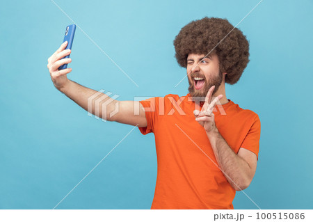 Portrait of man with Afro hairstyle wearing orange T-shirt showing v gesture, posing at phone camera, making selfie or having video call. Indoor studio shot isolated on blue background. Portrait of man with Afro hairstyle wearing orange T-shirt showing v gesture, posing at phone camera, making selfie or having video call. Indoor studio shot isolated on blue background. 100515086