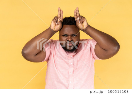 Portrait of man wearing pink shirt showing bull horn gesture, looking at camera with hostile angry expression, bully male threatening to attack. Indoor studio shot isolated on yellow background. 100515103