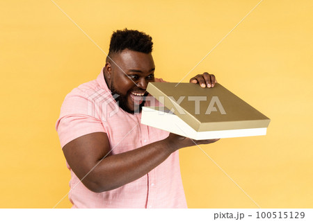 Portrait of optimistic man wearing pink shirt opening gift box and peeping inside with toothy smile, satisfied with present. Indoor studio shot isolated on yellow background. 100515129