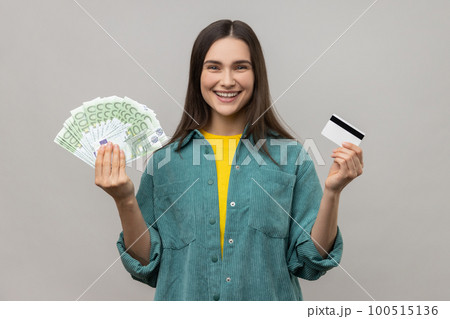 Happy satisfied young woman holding credit card and fan of euro cash looking at camera with toothy smile, wearing casual style jacket. Indoor studio shot isolated on gray background. Happy satisfied young woman holding credit card and fan of euro cash looking at camera with toothy smile, wearing casual style jacket. Indoor studio shot isolated on gray background. 100515136