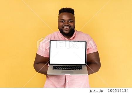 Portrait of happy smiling man wearing pink shirt showing laptop with blank display for advertisement, having satisfied expression. Indoor studio shot isolated on yellow background. 100515154