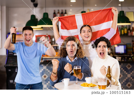 Company of young adult enthusiastic fans waving flag of Denmark and supporting national team with beer in pub 100515163