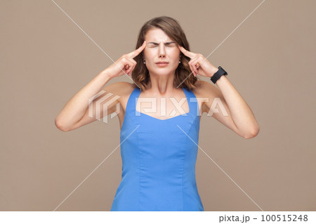 Sick unhealthy woman with wavy hair keeps fingers on temples, suffering terrible headache, standing with closed eyes, wearing blue dress. Indoor studio shot isolated on light brown background. 100515248
