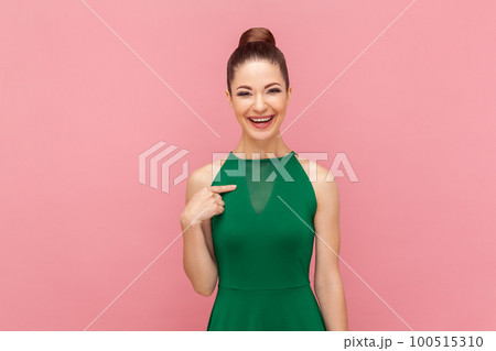 Portrait of smiling delighted attractive woman with bun hairstyle standing pointing at herself, being glad to be chosen, wearing green dress. Indoor studio shot isolated on pink background. 100515310