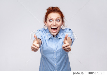 Portrait of positive redhead woman wearing blue shirt showing thumbs up, looking at camera with open mouth, surprised with excellent result. Indoor studio shot isolated on gray background. 100515328