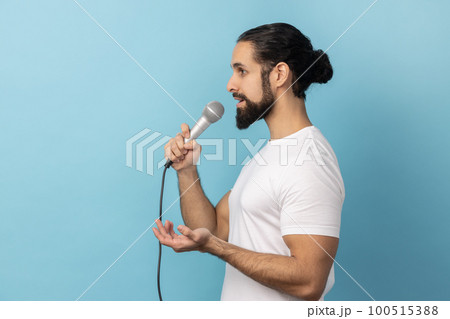 Side view of serious man with beard wearing white T-shirt, journalist holding microphone, discussing problems, reporting. Indoor studio shot isolated on blue background. 100515388