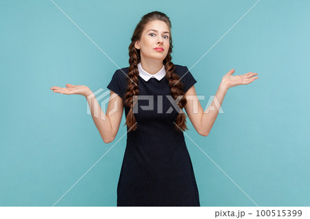 Puzzled uncertain young woman with braids spreads hands aside, shrugging shoulders, don't know how to make a decision, wearing black dress. woman Indoor studio shot isolated on blue background. 100515399