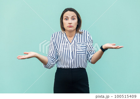Uncertain clueless attractive middle aged woman wearing striped shirt doesn't know correct answer, looking at camera, shrugging shoulders. Indoor studio shot isolated on light blue background. Uncertain clueless attractive middle aged woman wearing striped shirt doesn't know correct answer, looking at camera, shrugging shoulders. Indoor studio shot isolated on light blue background. 100515409
