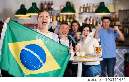 Company of emotional young adult fans supporting sports team of Brazil with state flag while resting with beer in bar 100515423