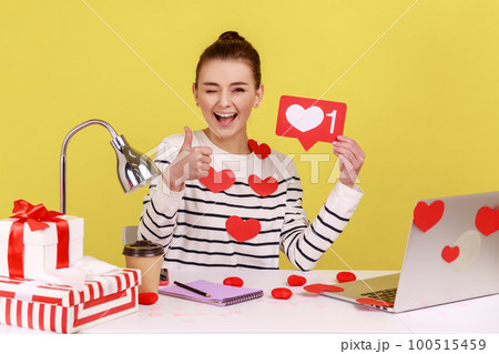 Smiling woman holding like counter template sitting at workplace covered heart sticks, making post in social network, showing thumb up. Indoor studio studio shot isolated on yellow background. Smiling woman holding like counter template sitting at workplace covered heart sticks, making post in social network, showing thumb up. Indoor studio studio shot isolated on yellow background. 100515459
