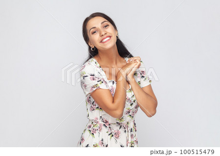 Portrait of cute charming beautiful woman wearing white dress looking at camera with toothy smile, keeps hands together, admires. Indoor studio shot isolated on gray background. Portrait of cute charming beautiful woman wearing white dress looking at camera with toothy smile, keeps hands together, admires. Indoor studio shot isolated on gray background. 100515479
