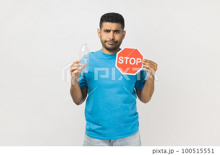 Portrait of sad serious unshaven man wearing blue T- shirt standing holding red stop symbol and empty plastic bottle, Earth protection. Indoor studio shot isolated on gray background. Portrait of sad serious unshaven man wearing blue T- shirt standing holding red stop symbol and empty plastic bottle, Earth protection. Indoor studio shot isolated on gray background. 100515551