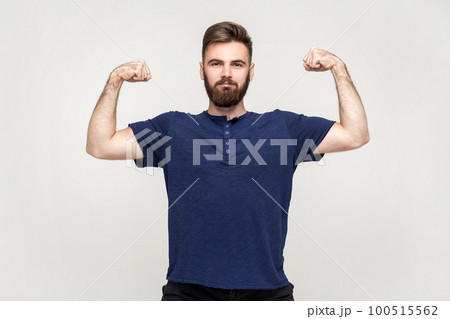Portrait of strong handsome man with beard wearing dark blue T-shirt looking assertive and raising arms to show biceps, feeling powerful. Indoor shot isolated on gray background. 100515562