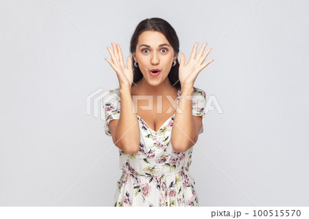 Portrait of astonished woman wearing white dress standing with mouth open in surprise, has shocked expression, hears unbelievable news, raised hands. Indoor studio shot isolated on gray background. Portrait of astonished woman wearing white dress standing with mouth open in surprise, has shocked expression, hears unbelievable news, raised hands. Indoor studio shot isolated on gray background. 100515570