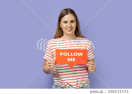 Portrait of pretty cute adorable blond woman blogger wearing striped T-shirt holding red card with follow me inscription, subscribe my vlog. Indoor studio shot isolated on purple background. 100515572