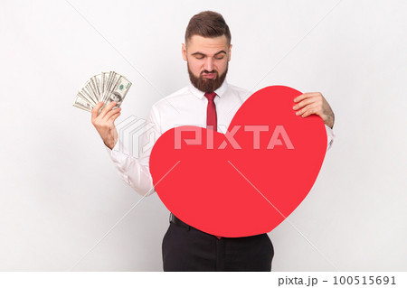 Portrait of young adult bearded man wearing white shirt and tie holding big dollar banknotes, looking at big red heart, choosing money, not love. Indoor shot isolated on gray background. Portrait of young adult bearded man wearing white shirt and tie holding big dollar banknotes, looking at big red heart, choosing money, not love. Indoor shot isolated on gray background. 100515691