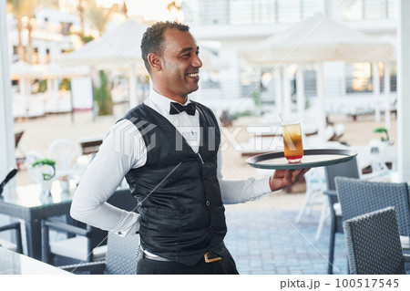 Black waiter in formal clothes is at his work outdoors at sunny daytime Black waiter in formal clothes is at his work outdoors at sunny daytime 100517545