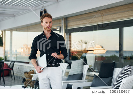 Holds laptop. Young man is outdoors at sunny daytime. Concept of vacation 100518497