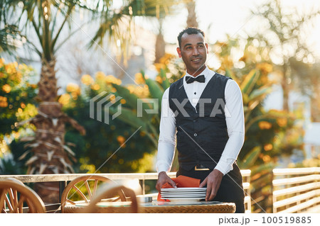 Wipes plates by red cloth. Black waiter in formal clothes is at his work outdoors at sunny daytime Wipes plates by red cloth. Black waiter in formal clothes is at his work outdoors at sunny daytime 100519885