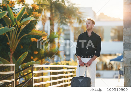 With travel bag. Young man is outdoors at sunny daytime. Concept of vacation 100519900