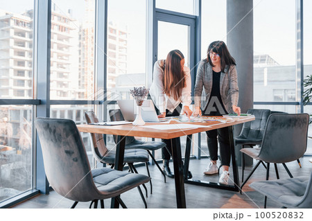 Standing and talking. Two women in formal clothes is indoors in the modern office works together 100520832